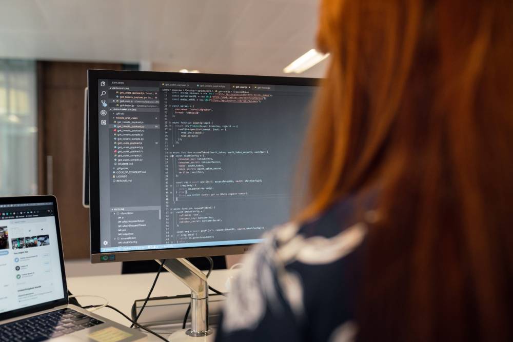 A programmer with long red hair views code on a large monitor, with a laptop displaying a social media page in the foreground.