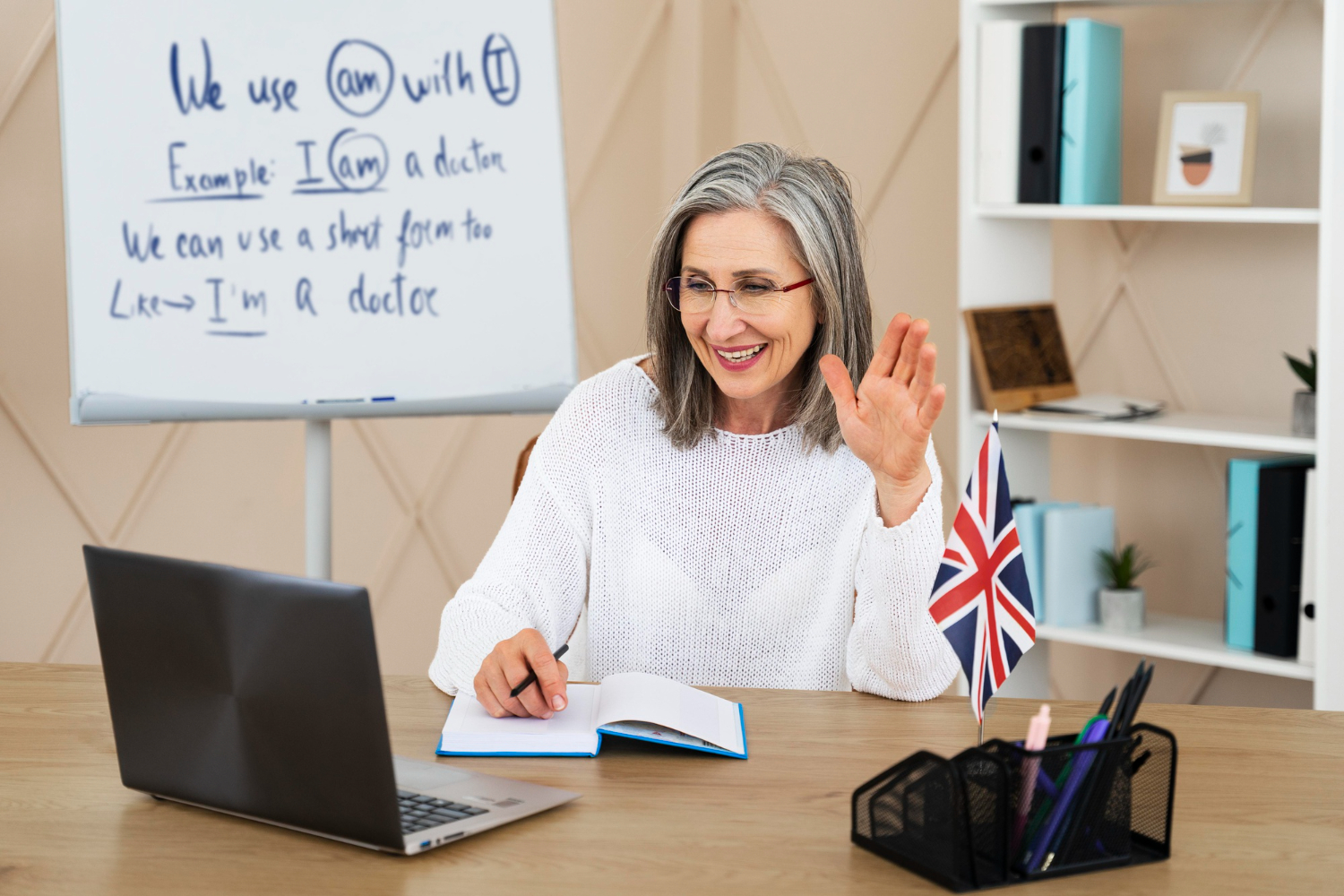 A woman in a white sweater gestures while sitting at a desk with a laptop, notebook, pens, and a small British flag, with a whiteboard behind her.