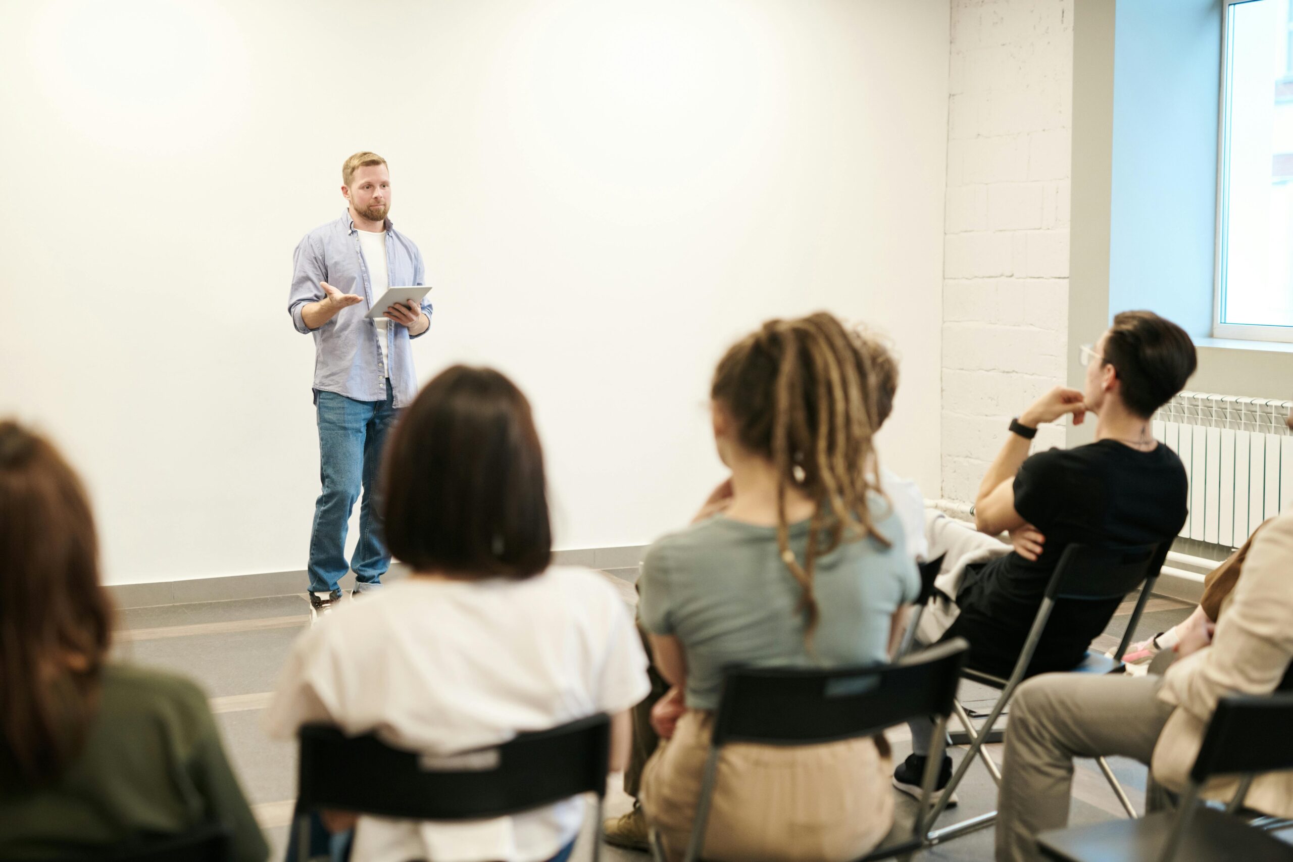 A speaker presents to an attentive audience seated in chairs, in a bright, minimalist room with white walls and a window.