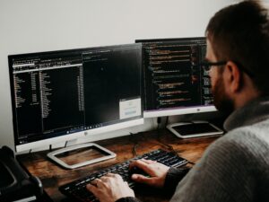 A person coding at a dual monitor setup, displaying file directories and programming scripts, with a dark wooden desk underneath.