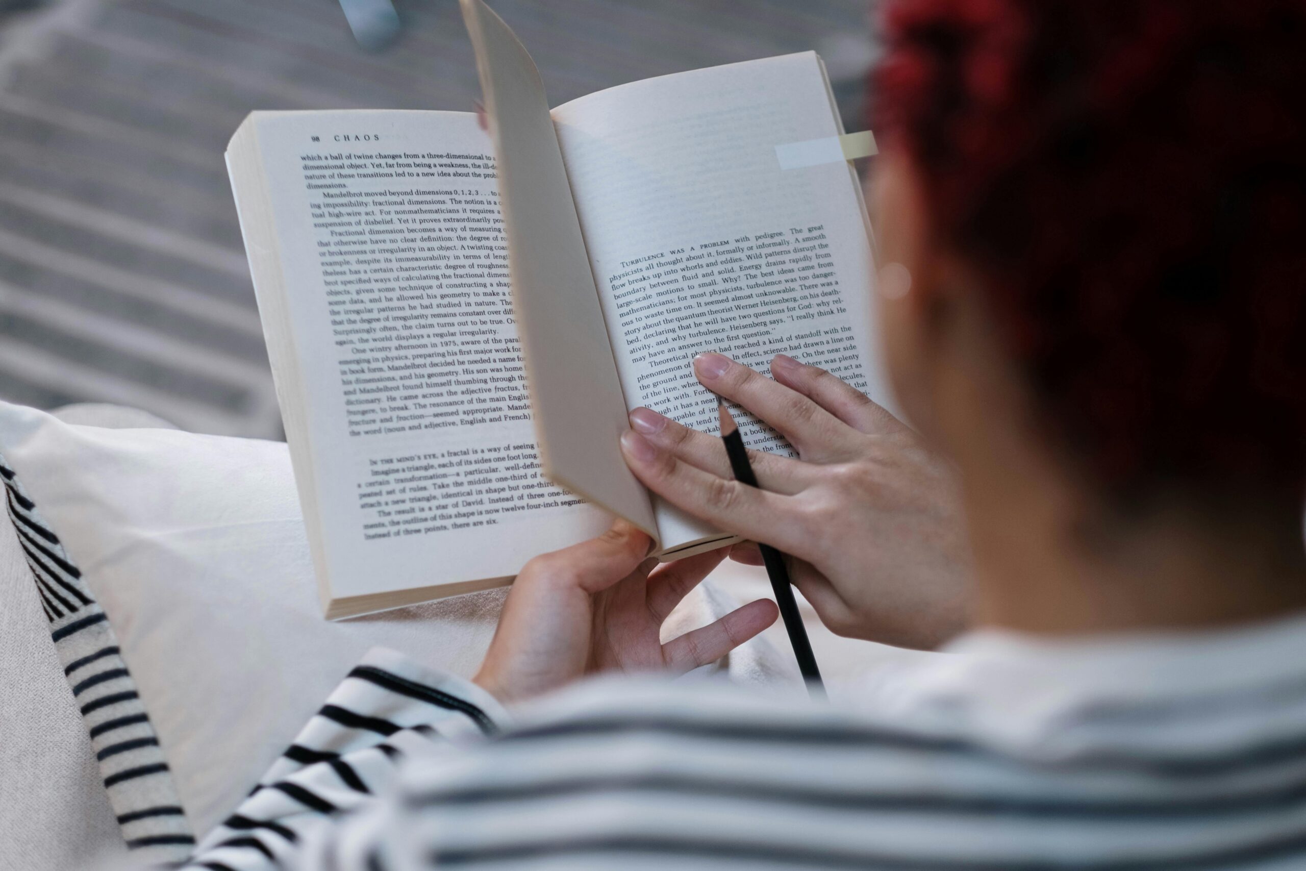 A person reads a book titled Chaos, holding it with one hand and using a pencil to take notes.