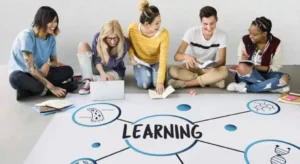 A group of students collaborate on a floor map with LEARNING at its center, surrounded by various educational symbols and materials.