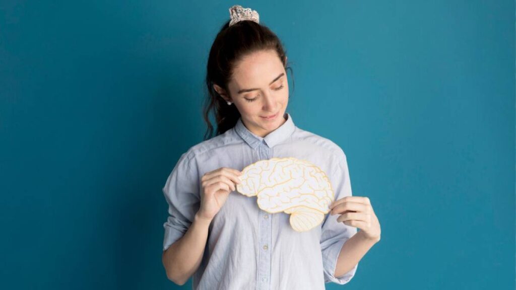 A person holds a paper cutout of a brain against a teal background, wearing a light blue button-up shirt.