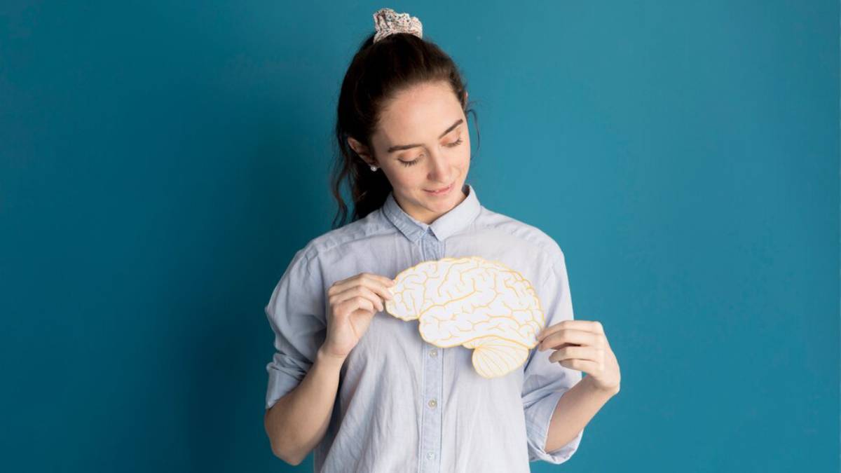 A person holds a paper cutout of a brain against a teal background, wearing a light blue button-up shirt.