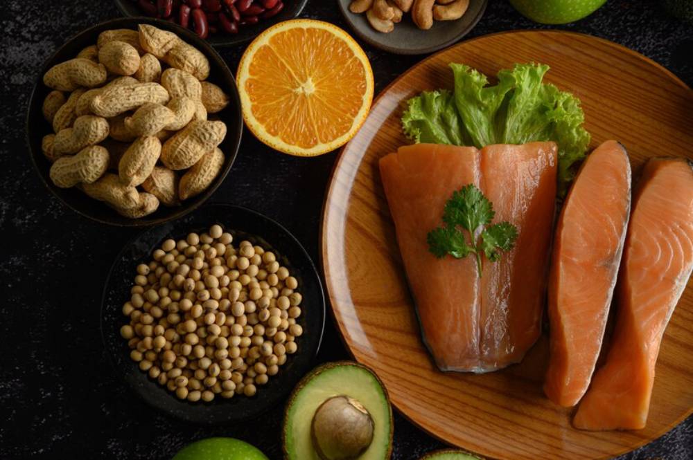 A wooden plate with salmon fillets topped with cilantro, surrounded by various nuts, seeds, orange slices, and green apple.