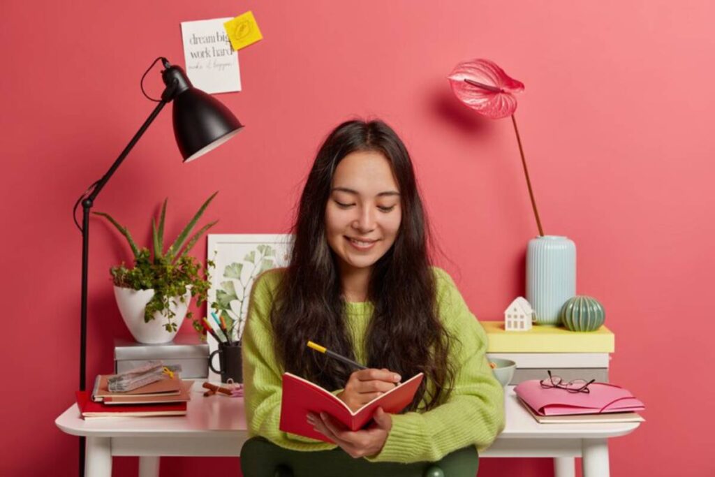 A person wearing a green sweater writes in a red notebook at a desk adorned with plants, books, and colorful decor against a pink wall.