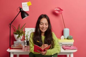 A person wearing a green sweater writes in a red notebook at a desk adorned with plants, books, and colorful decor against a pink wall.