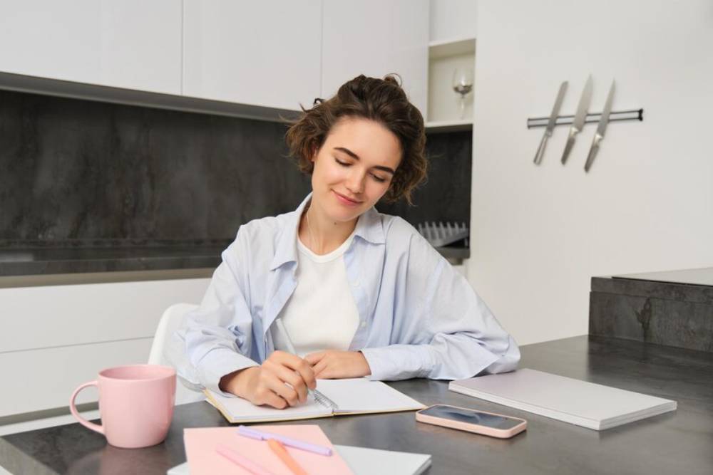 A person in a light blue shirt sits on table, writing notes in a notebook, with a pink mug and stationery nearby.