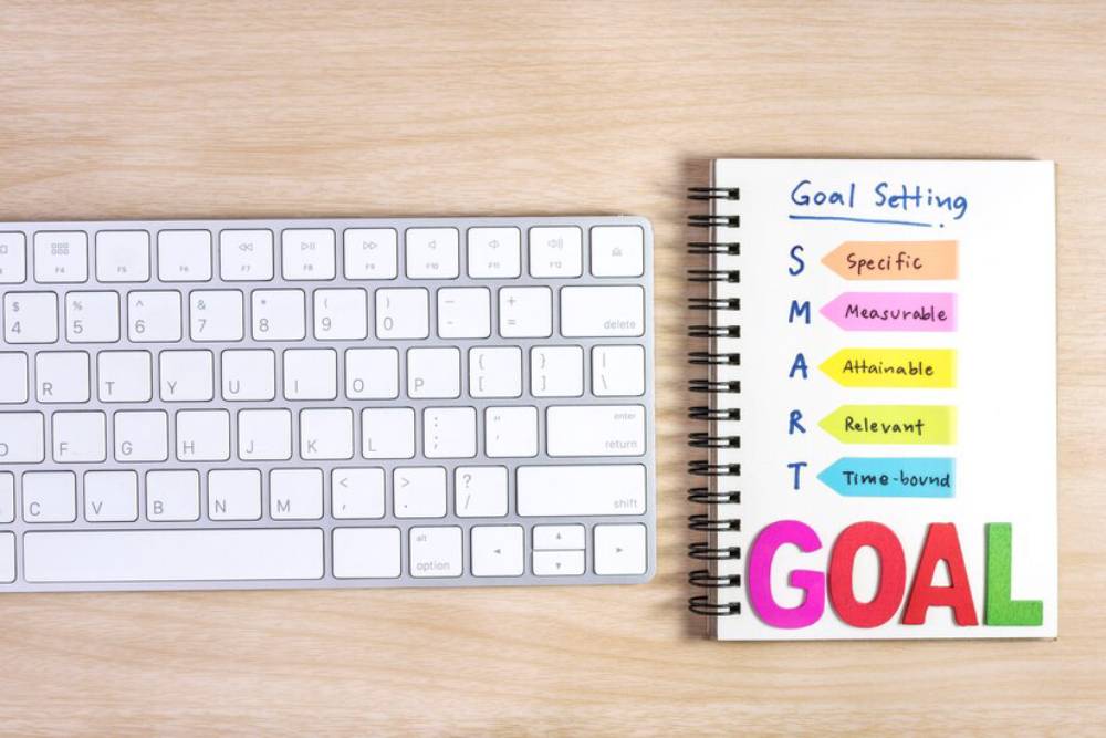 A white keyboard beside a spiral notebook, featuring colorful SMART goal-setting criteria: Specific, Measurable, Attainable, Relevant, Time-bound.