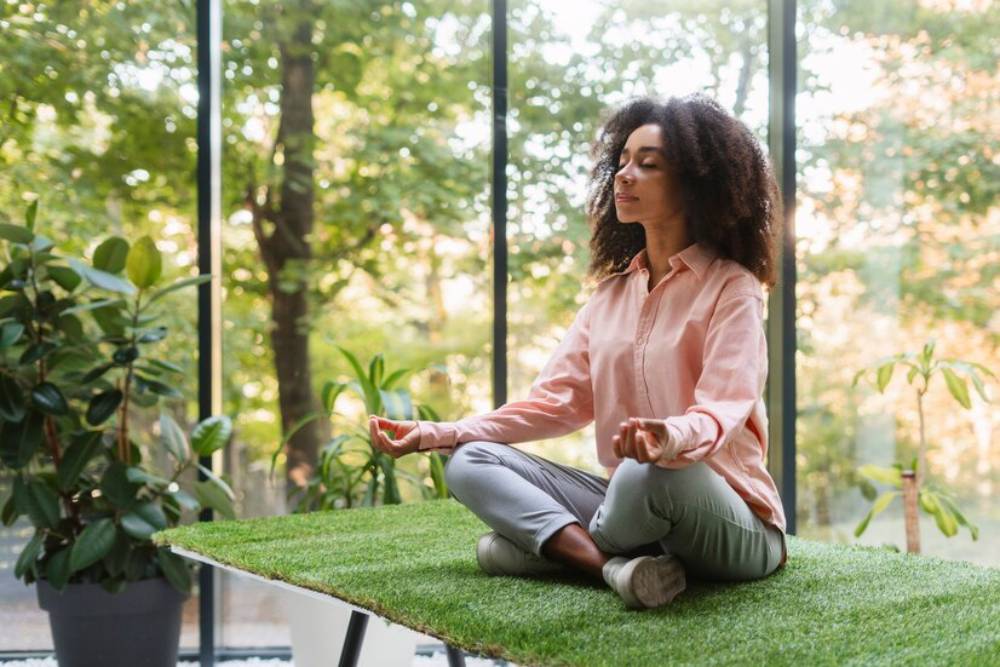 A woman sits cross-legged on a grassy surface indoors, meditating with plants, nature-filled setting.