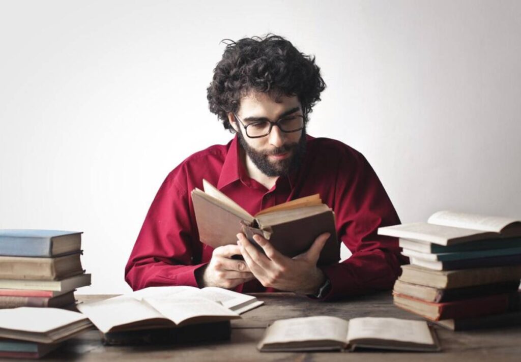 A polymat holds an open book, surrounded by stacks of books on a wooden table, immersed in learning.
