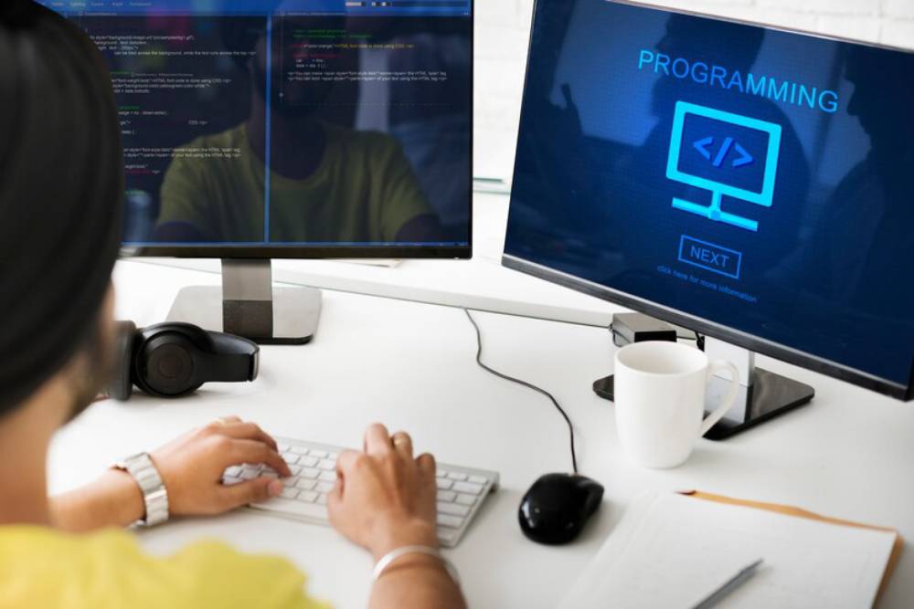 A person in a yellow shirt coding on a keyboard, with two monitors displaying programming content and a mug nearby.
