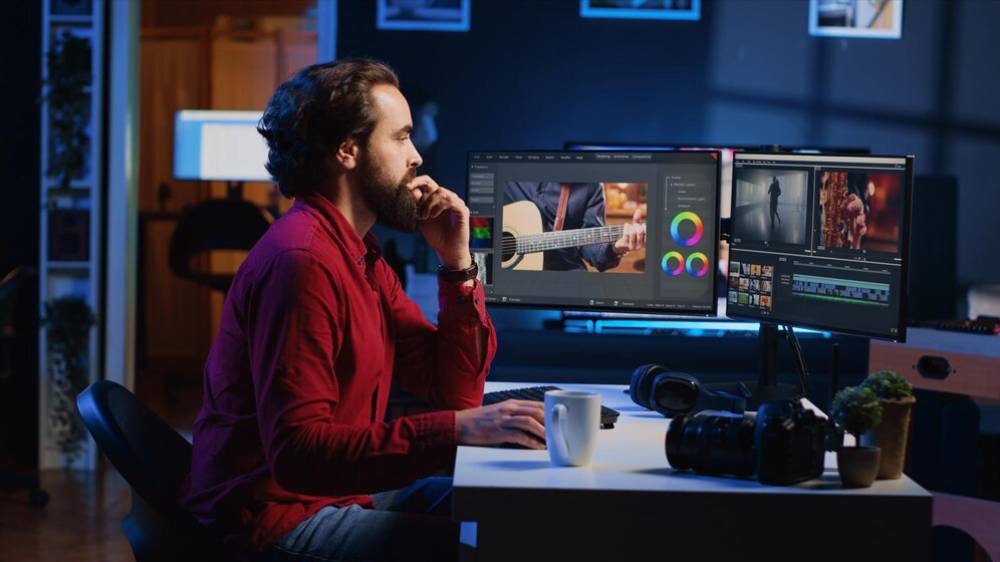 A person in a red shirt edits video content on dual monitors in a dimly lit workspace, surrounded by a coffee cup and camera gear.