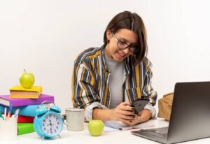 A person in a striped shirt engaged with their smartphone at a study desk, surrounded by books, a clock, and an apple.